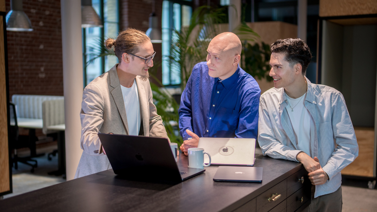 Drei Männer stehen in einem modernen Büro gemeinsam an einem hohen Tisch und unterhalten sich freundlich. Vor ihnen liegen zwei geöffnete Laptops und eine Kaffeetasse. Der Mann links trägt einen beigen Blazer, der in der Mitte ein blaues Hemd, und der rechts ein helles, gestreiftes Hemd. Im Hintergrund sind Pflanzen, große Fenster und Sitznischen zu sehen – die Atmosphäre wirkt konzentriert und kollegial.